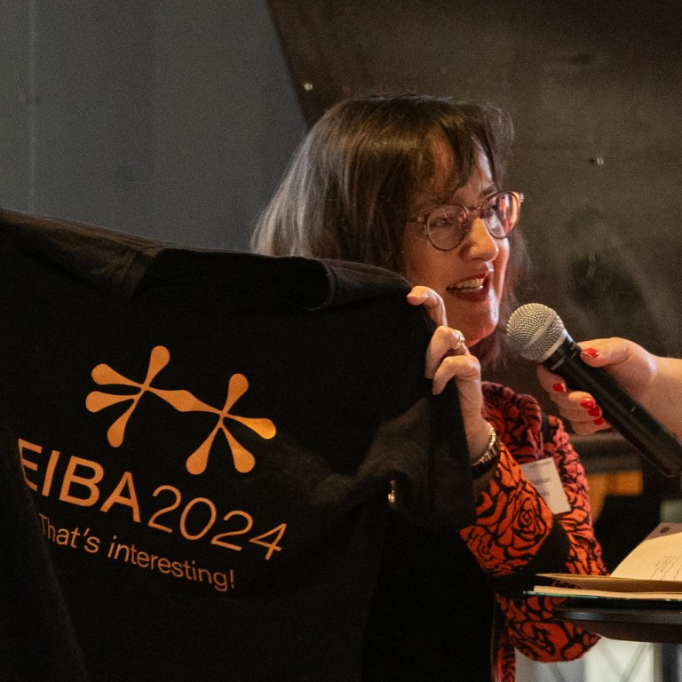 Rebecca Piekkari pitämässä kädessään mustaa t-paitaa, jossa lukeaa EIBA 2024-kongressin nimi.
