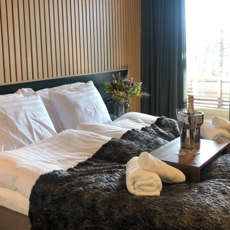 A double bed with towels on the bed and sparkling wine on a tray in a cooler and two glasses.