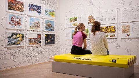 Teenagers sitting at the bench in the Mauri Kunnas Exhibition and looking at the exhibited drawings.