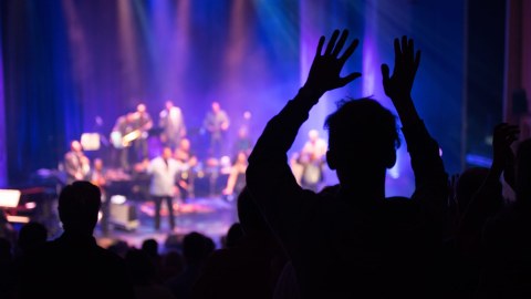 Taustalla lavalla esiintyy yhtye. Yleisö seisoo ja osoittaa suosiota kädet ylhäällä.