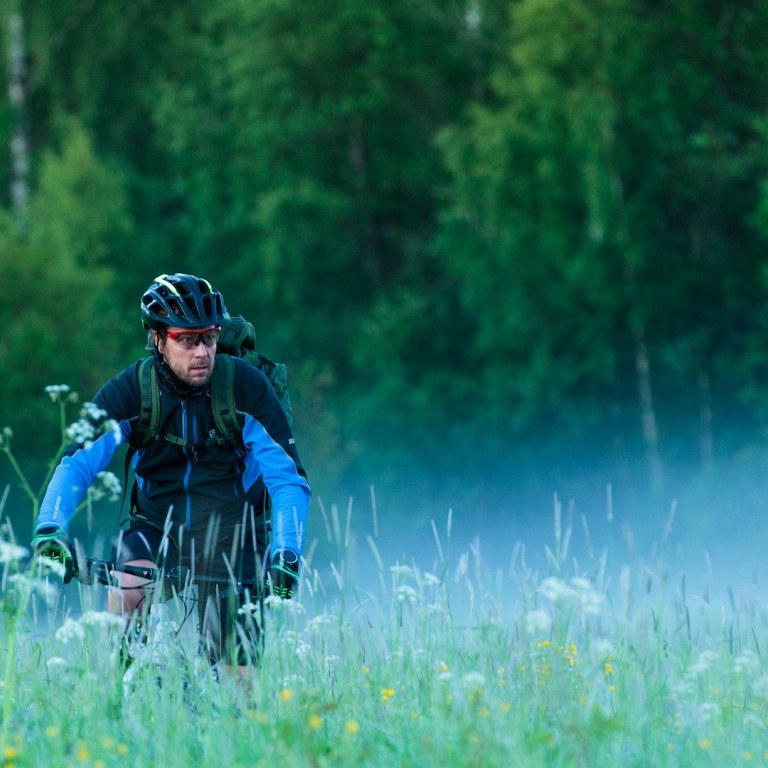 Mies pyöräilee hieman sumuisessa niittymaisemassa.