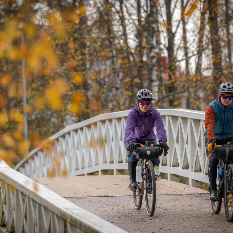 Kaksi pyöräilijää tunnelmallisella sillalla ympärillään ruskan värit.