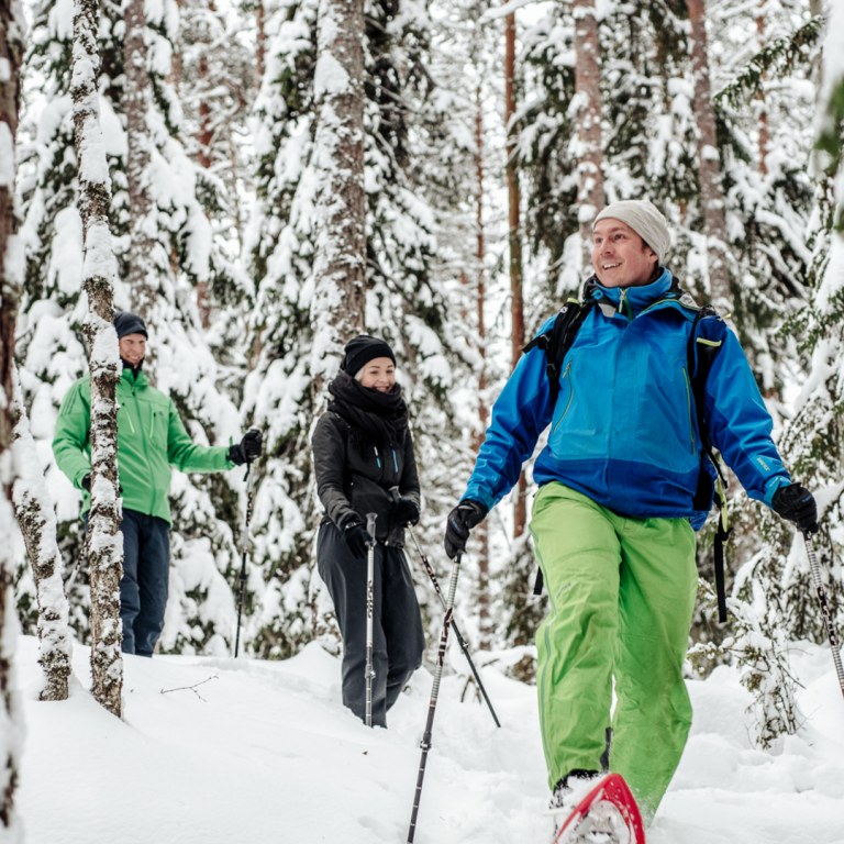 Iloisia lumikenkäijöitä kävelemässä ryhmänä Nuuksion lumisessa metsässä.