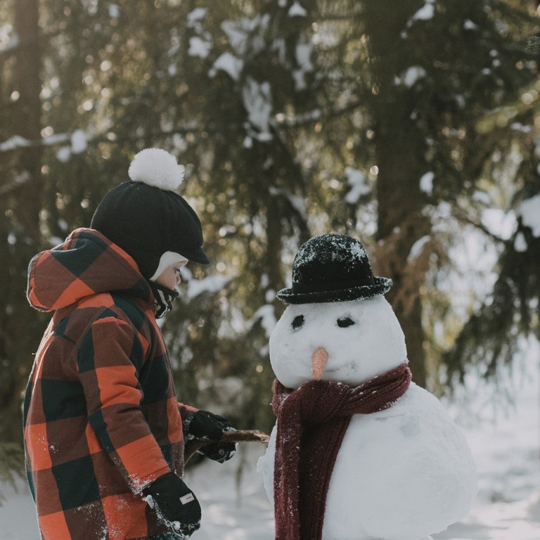 Lapsi seisoo lumiukon vieressä viimeistelemässä taideteosta lumisessa metsämaisemassa.