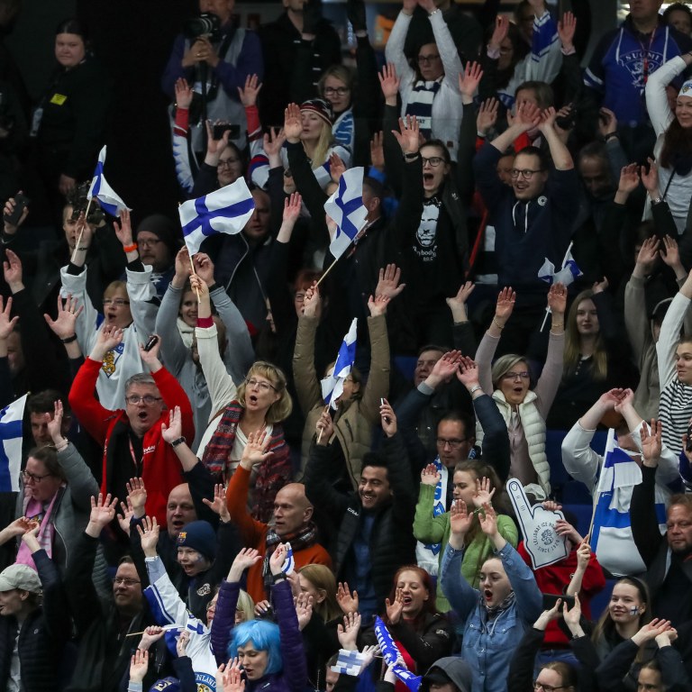 Kuva urheilukatsomosta, jossa ihmiset hurraavat ja kannustavat kädet ylhäällä ja suomenlippuja käsissään.