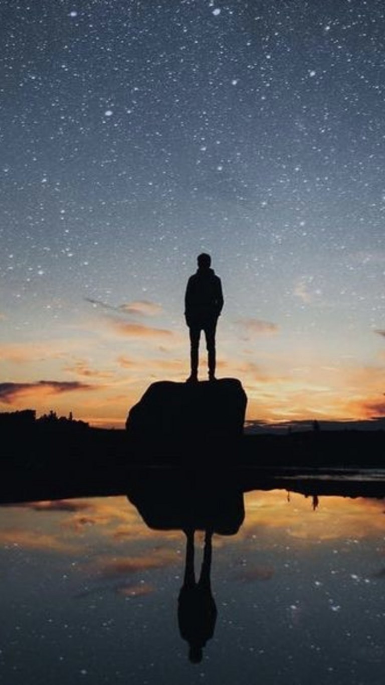 A person standing on a rock on the beach and looking at the dark starry sky.