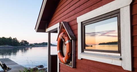 Puinen punainen saunarakennus meren rannalla, jonka ikkunasta heijastuu kesäinen auringonlasku.