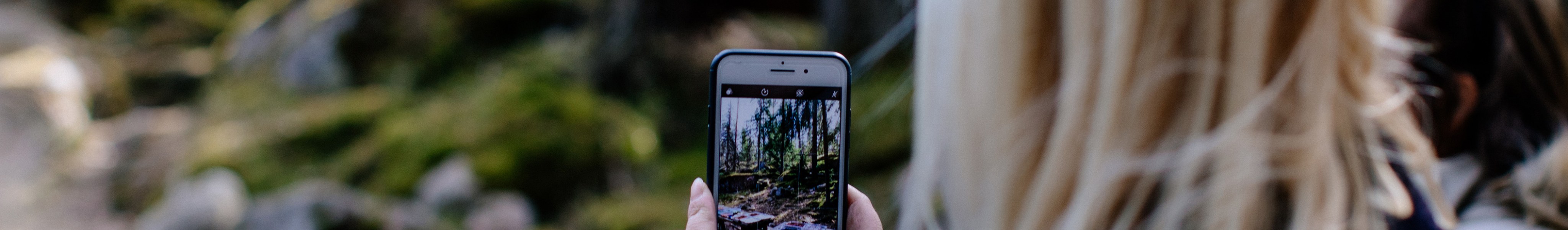 Nainen selin katsojaan kuvaamassa kännykkäkameralla edessään olevaa metsää.