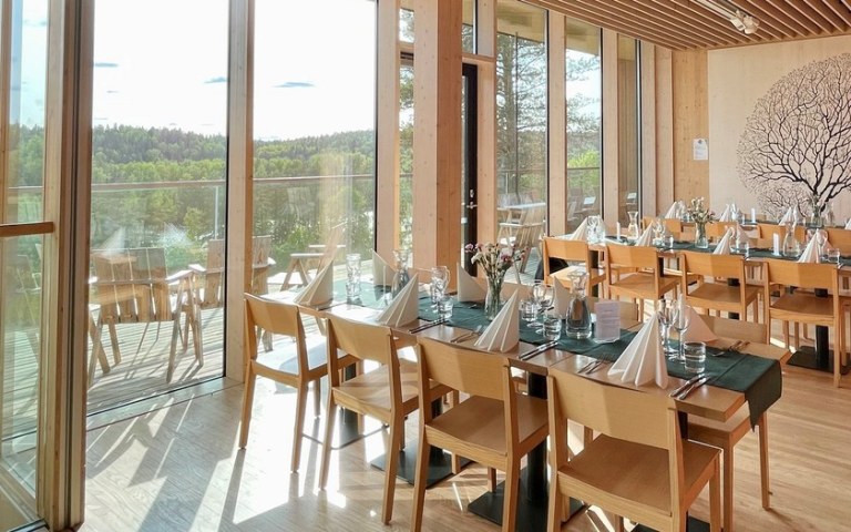 Two long wooden dining tables covered for dining. Tables in front of large windows with a view of the lake and the forest.