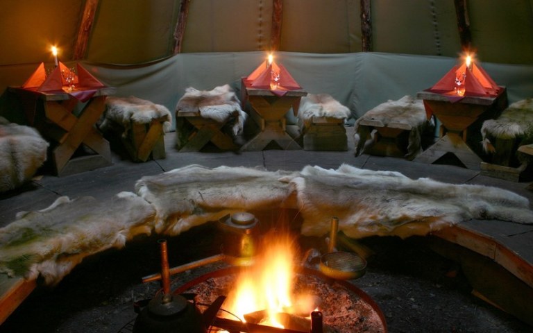A hut with reindeer harnesses on the long benches of the dining tables. Campfire in the middle.